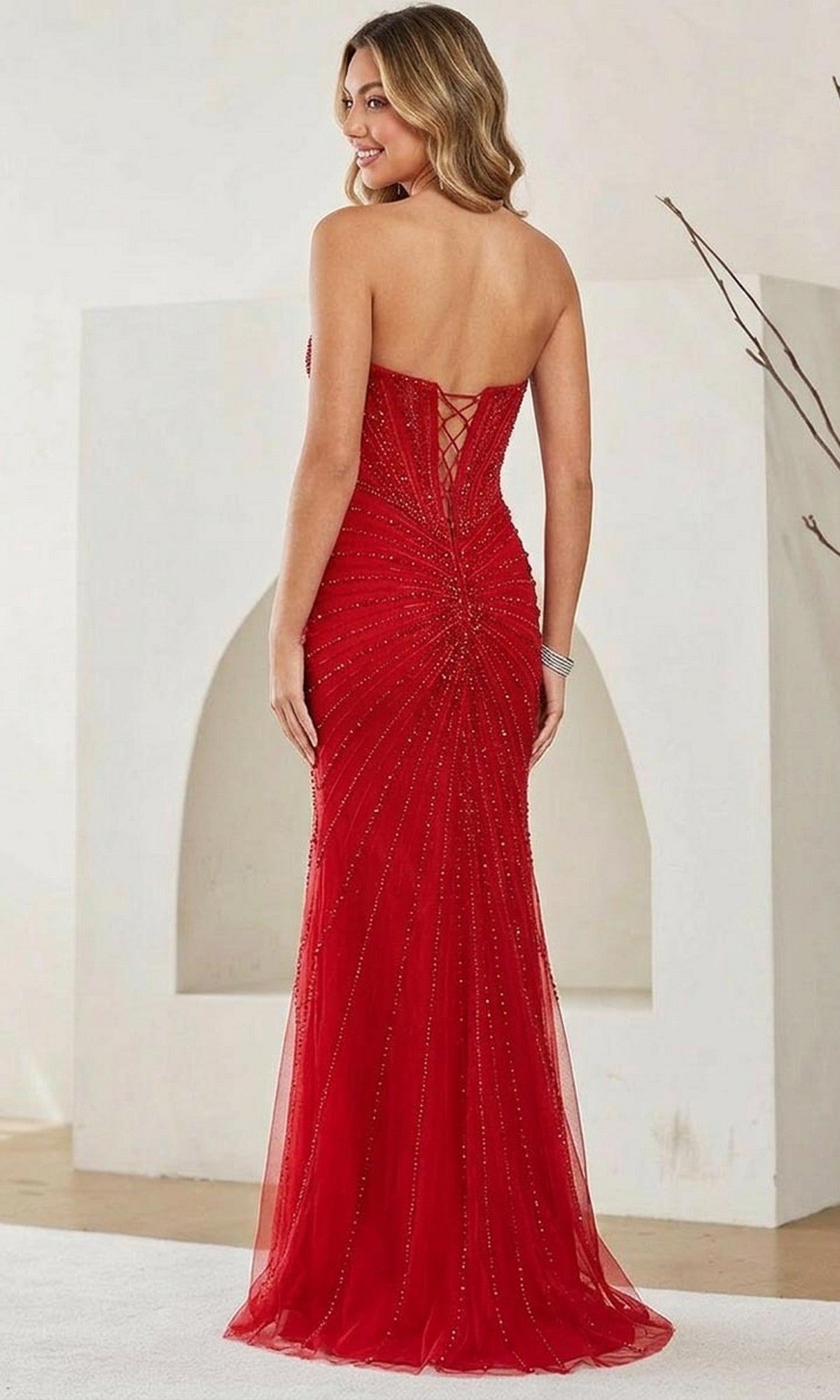 A woman wearing the Amelia Couture 9096 Long Prom Dress, a strapless beaded gown with a lace-up back, stands facing away in a minimalist indoor setting.